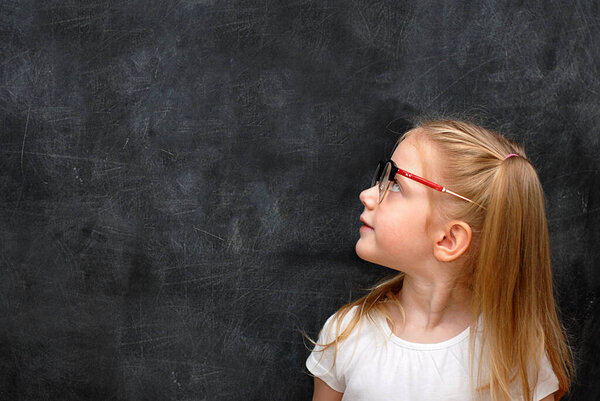 A blonde schoolgirl girl in glasses on a black background of a chalk board looks at the blackboard. Copy space.