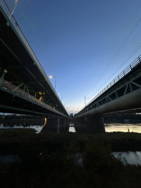 Varşova, Polonya - 24 Eylül 2025: Alacakaranlıkta nehirden geçen iki köprü, yapısal elementleri ve sakin su yansımalarını vurguluyor