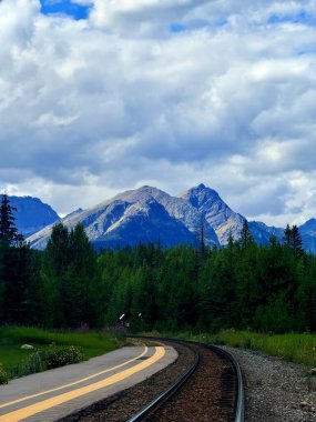 Sarmal demiryolu kıvrımları canlı yeşil ağaçların arasından geçiyor, arka planda yüksek dağlar ve dinamik bir gökyüzü, sakin bir açık hava manzarası yaratıyor.