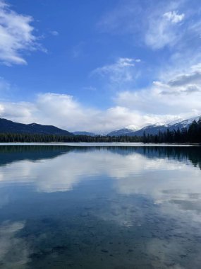 Dağların ve bulutların net yansımalarıyla sakin bir göl manzarası, yoğun ormanlar tarafından çerçevelenmiş, sakin ve huzurlu bir doğal ortam yaratıyor.