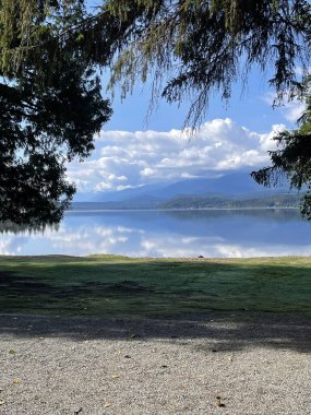 Berrak mavi gökyüzü ve yumuşak bulutların sakin su yüzeyinde yansıdığı bereketli ağaçlar ve dağların oluşturduğu sakin göl manzarası sakin bir atmosfer yaratıyor.