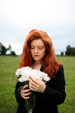 Açık bir alanda elinde bir buket beyaz çiçek tutan genç kızıl saçlı kadının sakin bir portresi, iç gözlem ve doğayla bağlantıyı çağrıştırıyor. Yumuşak ışık ve onun nazik ifadesi duygusal bir yansıma ve içsel esneklik duygusunu arttırır.