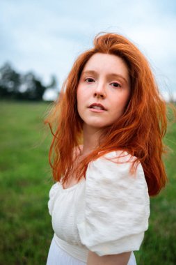 Natural light portrait of a red haired woman standing in a green field, wearing a white dress. The image evokes nostalgia, mental wellbeing, and quiet femininity within a peaceful rural landscape