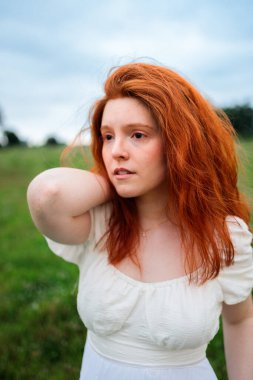Çimenli bir çayırda duran beyaz elbiseli genç kızıl saçlı bir kadının yakın portresi. Yumuşak doğal ışık ve rahat duruş özgünlük, içgözlem ve modern yaşam tarzı estetiğini iletir.
