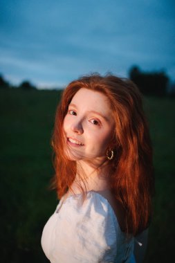 Mavi saatte yeşil bir tarlada duran beyaz elbiseli, kırmızı saçlı bir kadının portresini yakından çek. Yüzündeki ılık ışık, soğuk gökyüzü ile çelişiyor. Duygusal özgürlüğü, güveni ve iç huzuru simgeliyor.