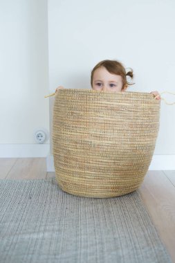 İki at kuyruklu, sarı taytlı ve bej tişörtlü küçük bir kız evde nesneler, oyuncaklar, hasır bir sepette saklanıyor, yatakta oyunlar oynuyor, parlak beyaz bir daire, çiçekler çiziyor.