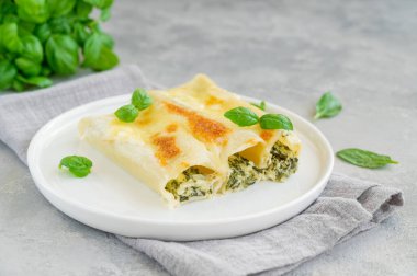 Geleneksel İtalyan cannellonisi, ricotta ve beşamel soslu ıspanak ve gri beton arka planda beyaz bir tabakta taze fesleğen yaprağı. Vejetaryen yemeği. Boşluğu kopyala
