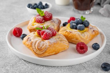 Danish puff pastry cakes with custard cream and fresh berries, sprinkled with powdered sugar on a white plate on a concrete background