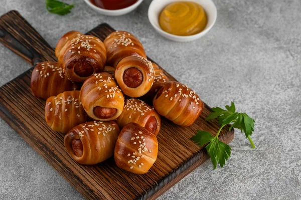 Mini sosisli, hardal ve ketçaplı. Maya hamuru kapağında pişmiş sosisler. Battaniyedeki domuzlar gri arka planda tahta bir tahtanın üzerinde. Üst görünüm, alanı kopyala