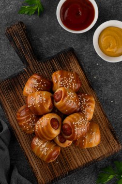 Mini hot dog ısırıkları. Maya hamuru kapağında pişmiş sosisler. Ahşap bir tahtanın üzerinde, koyu beton bir arka planda, battaniyedeki domuzlar. Üst görünüm