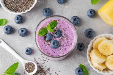 Yaban mersinli meyve suyu, muz, chia tohumu ve gri beton arka planda bir bardakta yoğurt. Sağlıklı yemek.