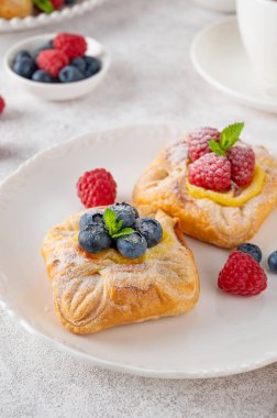 Danish puff pastry cakes with custard cream and fresh berries, sprinkled with powdered sugar on a white plate on a concrete background