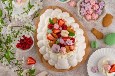 Paskalya yumurtası şeklinde krem şantili Pavlova pastası, üzeri taze böğürtlen ve beton bir arka planda tahta üzerinde çeşitli tatlılar ile kaplı. Üst görünüm, alanı kopyala