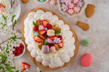 Paskalya yumurtası şeklinde krem şantili Pavlova pastası, üzeri taze böğürtlen ve beton bir arka planda tahta üzerinde çeşitli tatlılar ile kaplı. Üst görünüm, alanı kopyala