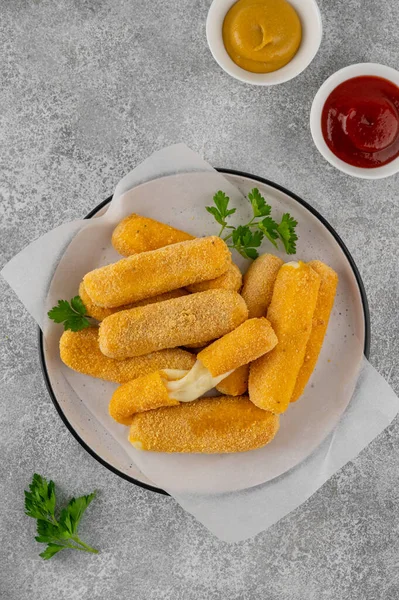 Ketçap ve hardal sosuyla servis edilen maydanozlu bir tabakta kızarmış mozzarella çubukları. Altın rengi kızartılmış doku, gri arka planda iştah açıcı aperatif..