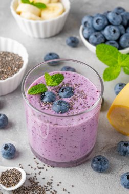 Yaban mersinli meyve suyu, muz, chia tohumu ve gri beton arka planda bir bardakta yoğurt. Sağlıklı yemek.