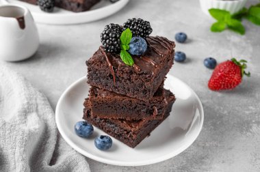 Çikolata kaplı bir yığın browni, gri beton arka planda beyaz bir tabakta taze böğürtlen ve nane yaprakları. Tatlı nefis.