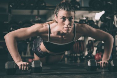 Spor salonunda dambıllarla egzersiz yapan genç bir kadın.