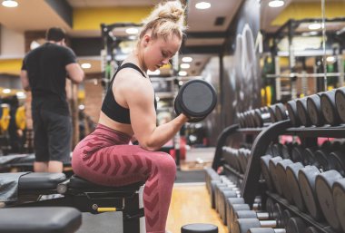 Spor salonunda dambıllarla egzersiz yapan genç bir kadın.