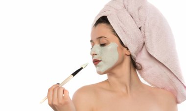 Yüzünde havlu olan güzel genç bir kadın beyaz arka planda izole edilmiş kafasına kozmetik maske takıyor.