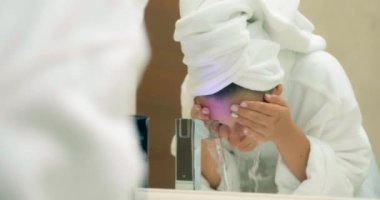 Bornozlu kadın banyodaki büyük aynaya bakarak yüzünü yıkıyor.