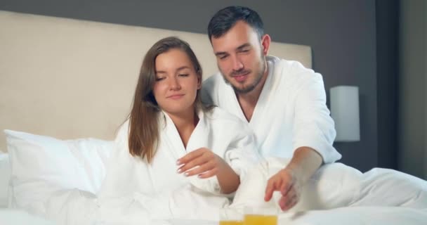 Pareja en albornoces beber jugo de naranja acostado en una cama grande — Vídeos de Stock