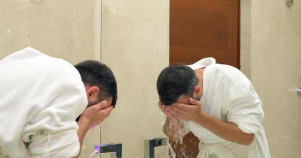 Hombre con barba en albornoz blanco lava la cara en el baño — Vídeo de stock