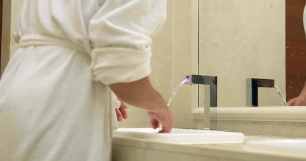 Mężczyzna w szlafroku naciska przycisk, aby uruchomić wodę i myje twarz — Wideo stockowe