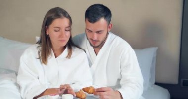 Harika bir çift, büyük yatakta kahvaltı yapan kruvasanlar yiyor.