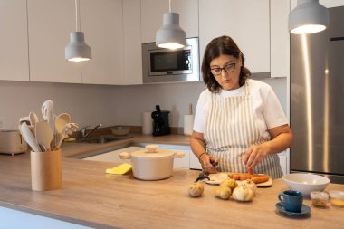 Modern bir mutfakta evde yemek pişirirken önlük takan, havuç ve patates doğrayan olgun bir kadın. Günlük yaşam tarzı, sağlıklı yemek hazırlama, sıcak ışık ve fotokopi alanı olan gerçek bir ev sahnesi..