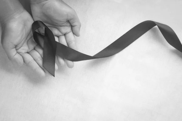 Hand holding Black ribbon on white fabric background with copy space, symbol of Skin Cancer awareness month on May, Melanoma cancer, Mourning ribbon symbolic. Healthcare medical and insurance concept.
