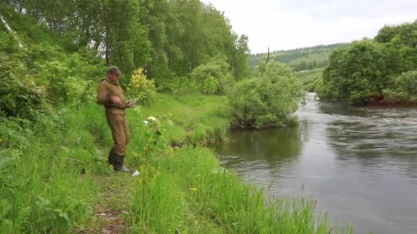 A man in a protective suit of protective color against encephalitis mites and rubber boots caught a fish in the river with a fishing rod. Summer, fishing on the river in Siberia — Stock Video