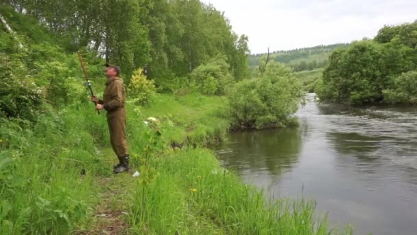 A man in a protective suit of protective color against encephalitis mites and rubber boots caught a fish in the river with a fishing rod. Summer, fishing on the river in Siberia — Stock Video