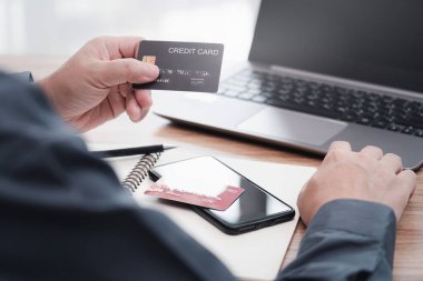 İşadamı internet bankacılığı yapıyor, ödeme yapıyor ya da internetten satın alıyor kredi kartı bilgilerini akıllı telefondan giriyor ve bilgisayarını masada çalışırken kullanıyor.