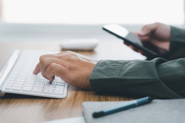 Ofiste çalışan akıllı telefonları tutan bir adamın masaüstü klavyesinde yazı yazması. 