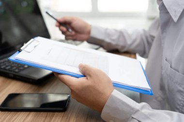 Kapatın. Bir adam grafik veri belgesini tutsun ve ofiste çalışan dizüstü bilgisayarı ve ahşap masada çalışan akıllı telefonu kullansın.