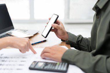 Mühendis teknisyeni iş yerinde meslektaşları ve dizüstü bilgisayarlı kalem hesap makinesi ve çizimleriyle ekranda akıllı telefon gösterdi