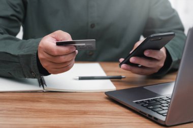 İşadamı internet bankacılığı yapıyor, ödeme yapıyor ya da internetten satın alıyor kredi kartı bilgilerini akıllı telefondan giriyor ve bilgisayarını masada çalışırken kullanıyor.