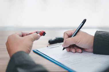 Araba sigortası kavramı: Adam kalem kullanıyor ahşap masaya sigorta belgeleri yazıyor ve elinde oyuncak bir araba modeli var