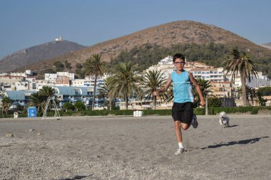 Sahilde koşan bir çocuk ve arkasında dağlar ve evler olan beyaz bir Malta bichon köpeği.