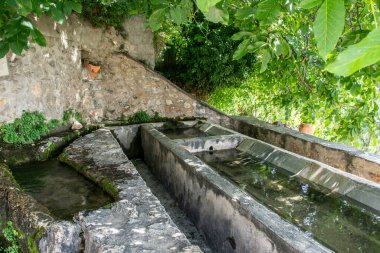 Yegen çıkışındaki taştan halk çamaşırları. Etrafı yeşilliklerle çevrili ve gölgede.