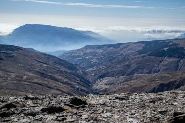 Önünde kayrak zemini, arkasında bir vadi ve arkasında alçak bulutlar denizi olan dağlar olan dağlık bir arazi.