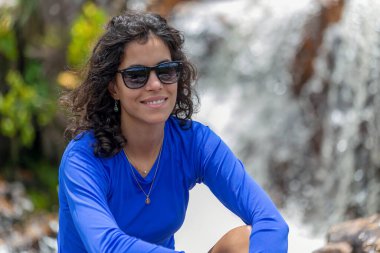 Esmer bir kadın şelalenin yanında oturuyor güneş gözlüğü ve mavi bir gömlek takıyor. Macera gezisi. Tatilde. Boş zaman.