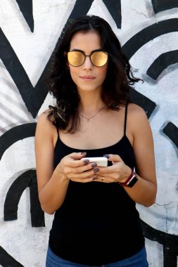 Güzel bir esmer kadın cep telefonunda mesajlaşıyor. Üzerinde de siyah tişört, mavi kot pantolon ve sarı aynalı gözlük var. Sao Paulo sokaklarında sabah güneşi. Moda.