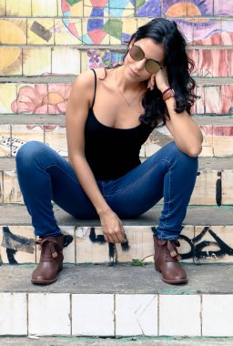 Basit siyah tişört, mavi kot pantolon, kahverengi bot ve aynalı güneş gözlüğü takan güzel esmer bir kadın merdivende oturuyor. So Paulo sokaklarında sabah güneşi. Moda. 