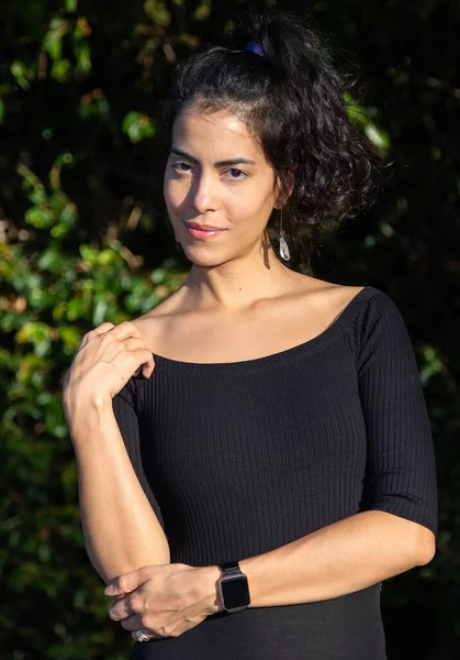 A Brazilian woman model photographed at dusk. She wears curly hair, bijouterie earrings and a black dress in the late afternoon. Brazilian model. Lifestyle. Fashion.