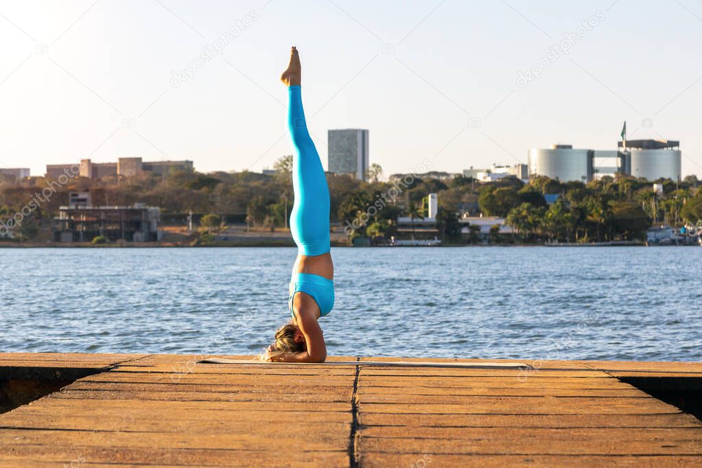 A blonde woman doing yoga pose on the edge of a lake at dusk. Body health. Lifestyle. In the background the city of Brasilia, Brazil.