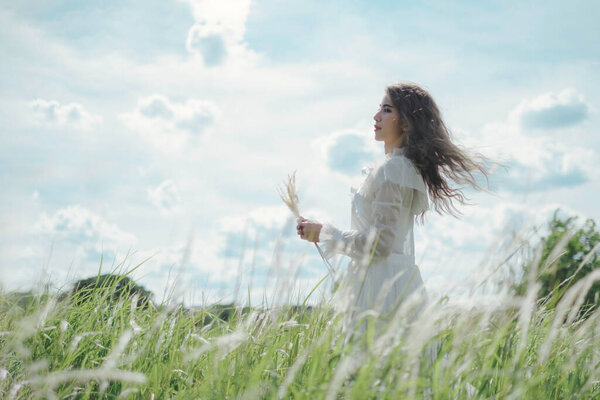 Young beautiful woman with brown spiral hair blowing by the wind of summer, holding fountain grass, looking away, standing in meadow or grass field.
