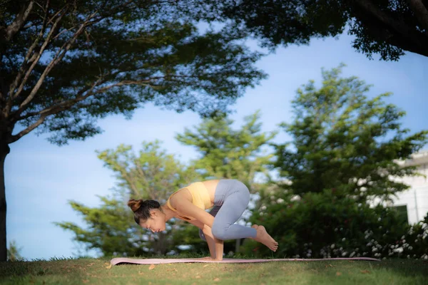 An asian woman in yellow sport bra and gray legging doing yoga in crow pose at, park under camber of big tree. Yoga practicing in nature, greenery, and freshness.