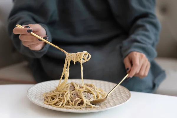 Woman hand with black colour nail paint rolling spaghetti with golden fork, blurred teal green sweater dress, looking delicious spaghetti carbonara on whithe plate and white table